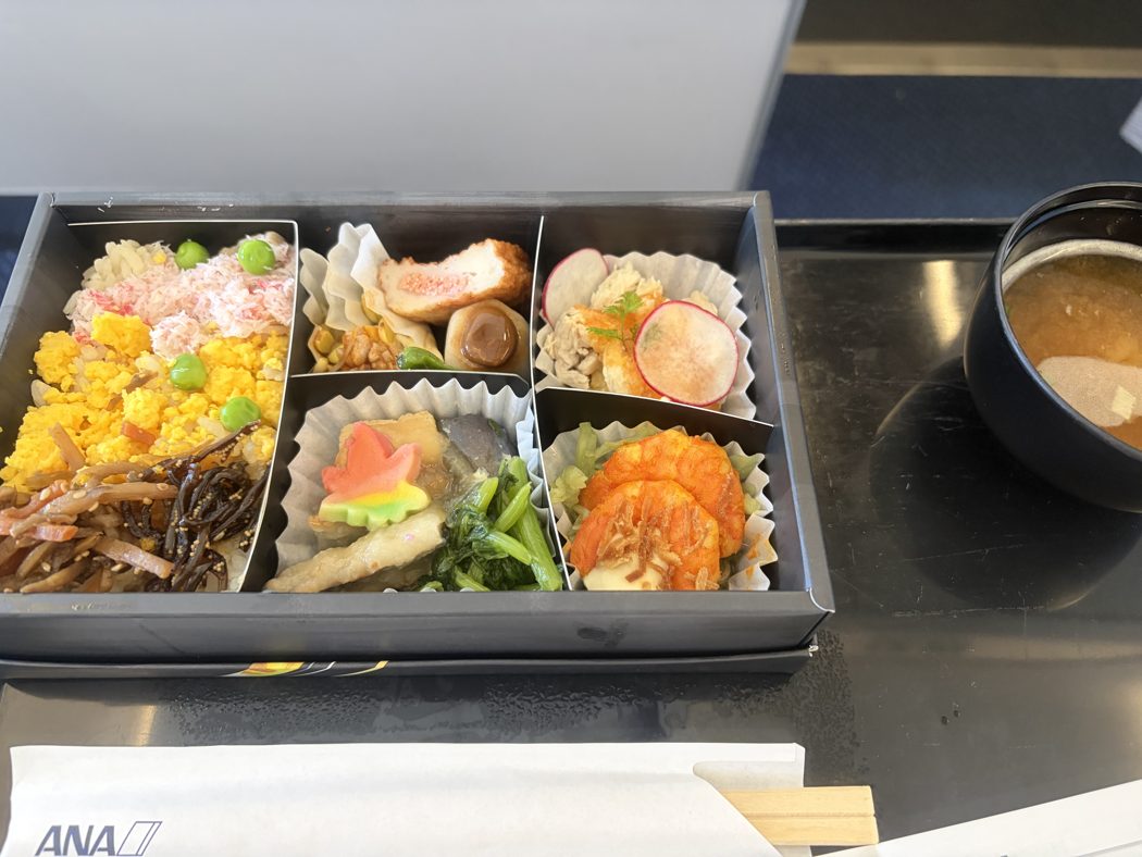 ANA国内線ファーストクラスの昼の機内食。仕切りのあるお弁当箱に錦糸卵とかにのご飯やおかずが入り、横に味噌汁のカップが置かれている