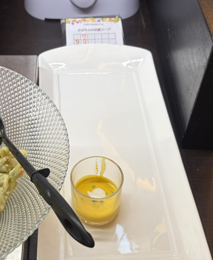 小さなグラスに注がれた冷製かぼちゃスープ。白い皿の上に置かれ、奥に料理名の札がある