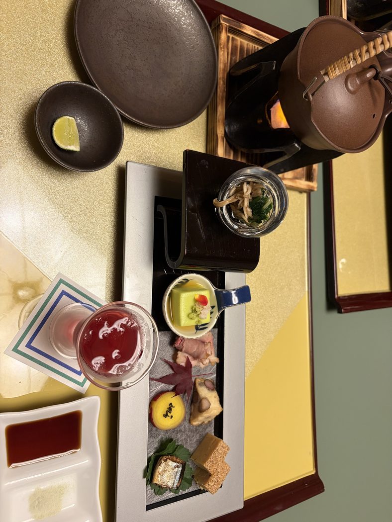 部屋で食べた朝食。長皿の盛り合わせ、緑色の豆腐の小鉢、赤い飲み物、奥に火にかけた鍋
