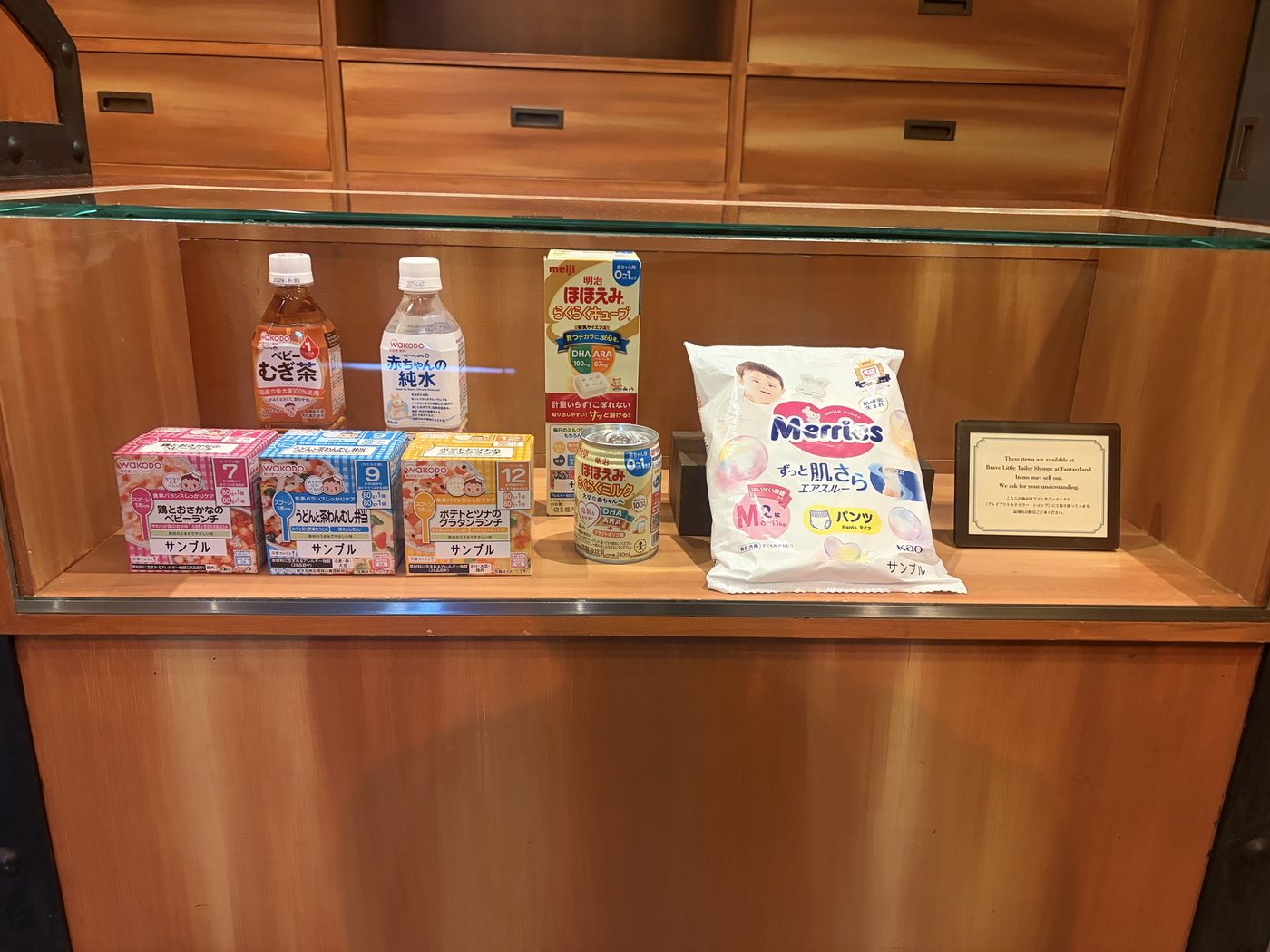 ベビーセンター内の展示ケース。和光堂のベビーむぎ茶と赤ちゃんの純水、栄養マルシェ3種、明治ほほえみらくらくキューブとらくらくミルク、メリーズのパンツMサイズ2枚入が並び、取り扱い店舗の案内板が添えられている
